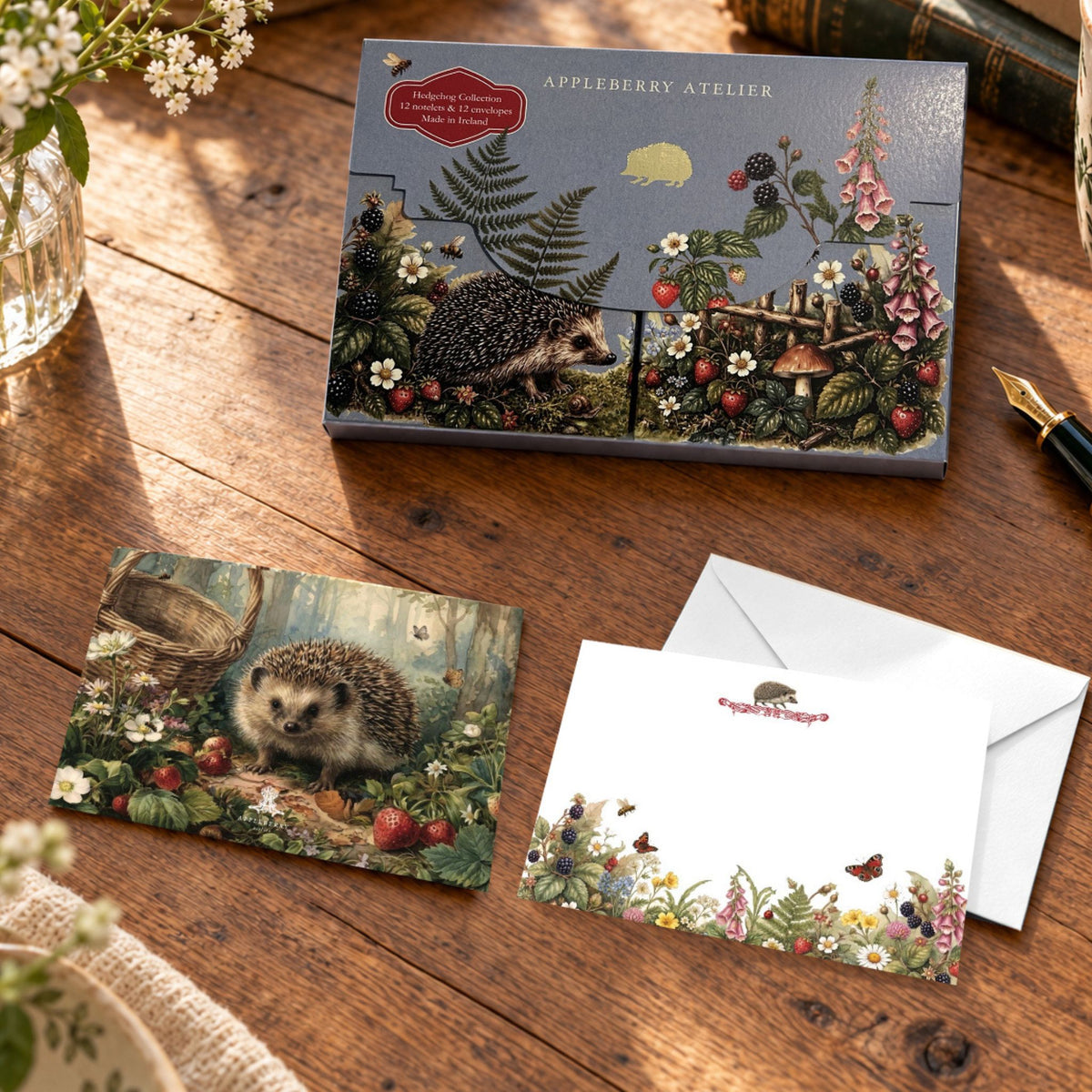 A styled flat-lay of notelet card and matching envelope on a rustic wooden table. The card features a detailed vintage-style illustration of a hedgehog in a forest, and the storage folder has a matching floral border.