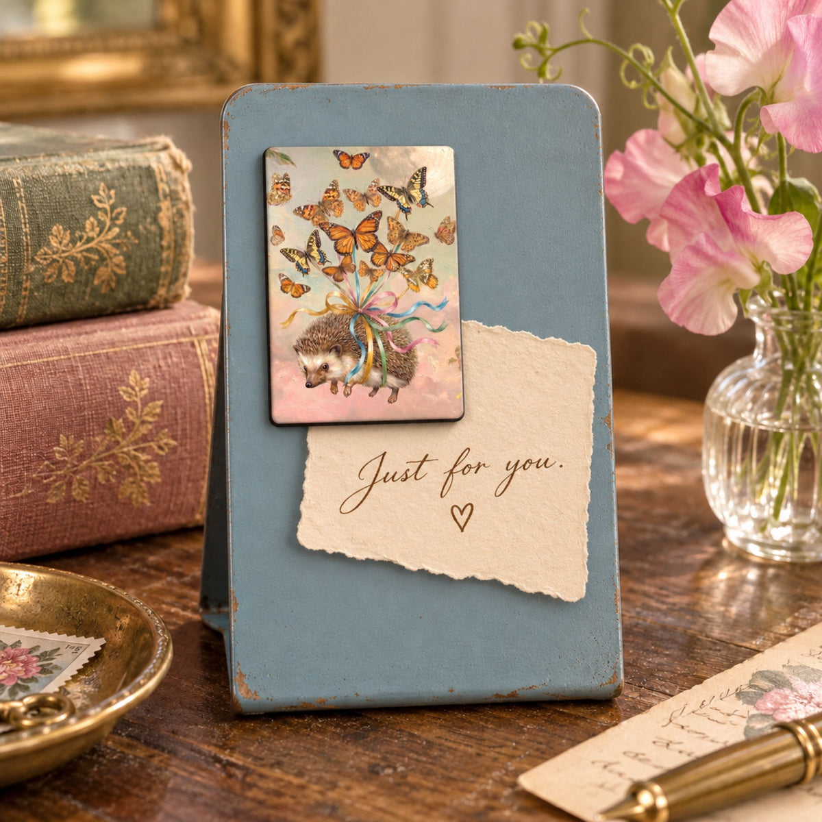 A small blue notice stand, with a hedgehog magnet displayed on a wooden table, featuring a butterfly-and-hedgehog illustration and the words 'Just for you.' with a torn paper note.