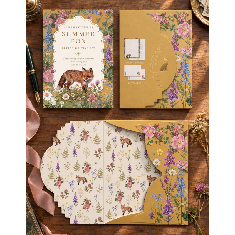 A decorative letter writing set displayed on a wooden surface, including a gold envelope with floral fox artwork, a patterned card set, and matching paper pieces arranged in a fan. The scene is styled with ribbon, a pen, and dried flowers.