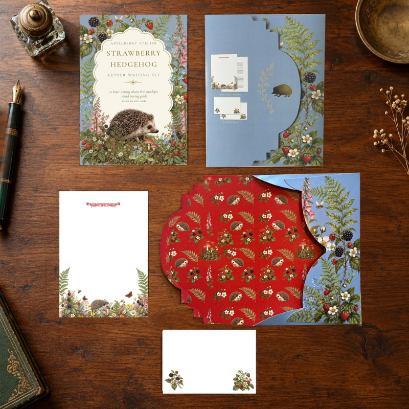A flat-lay presentation of a stationery suite for 'Strawberry Hedgehog' on a dark wooden table. The set includes a main invitation card with a hedgehog illustration, a blue envelope with floral borders, a white letterhead card, a red patterned envelope, and a small white card, styled with vintage props like a brass bottle stopper, a gold pen, and a brass bowl.