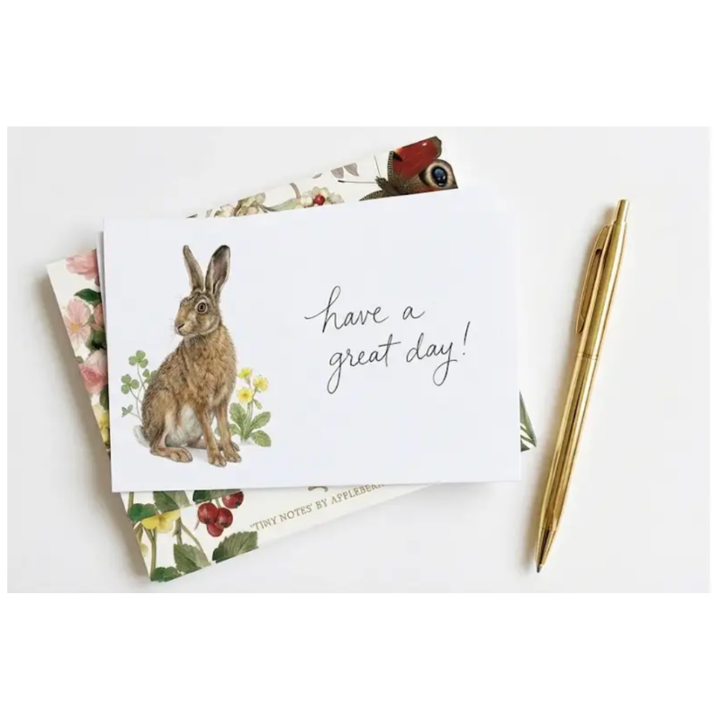 A white greeting card with a watercolor hare illustration and the handwritten text 'have a great day!' is styled on top of floral patterned cards, next to a gold pen on a white background.