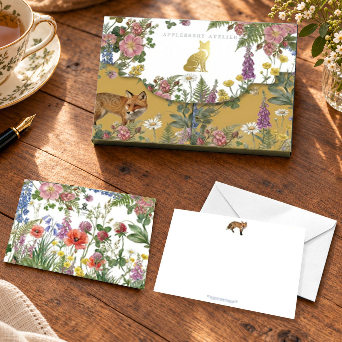 A boxed stationery set displayed on a wooden table, including a floral and fox-themed outer box, a matching notelet, an envelope, and a small note card. A teacup, gold pen, and flowers are arranged around it as styled props.