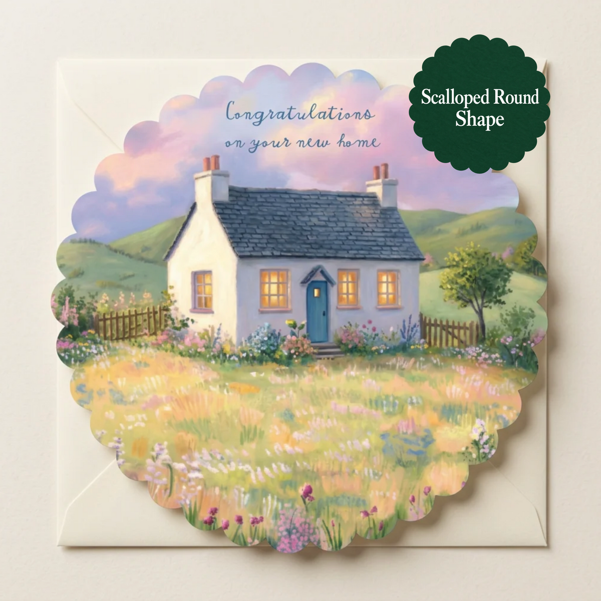 A scalloped round greeting card featuring a painted cottage scene with flowers and hills, shown on a white envelope. The card includes the text 'Congratulations on your new home' and a dark green badge that reads 'Scalloped Round Shape.