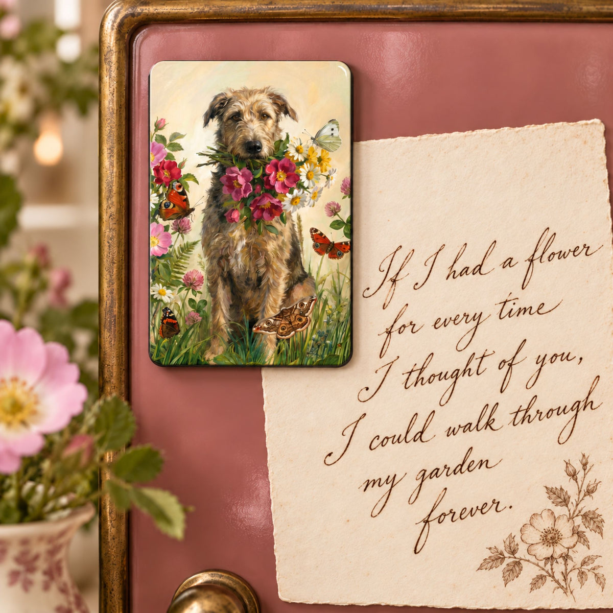 A rectangular printed magnet featuring a scruffy dog surrounded by colourful flowers and butterflies, attached to a pink surface next to a handwritten note. The scene is styled with flowers and a wooden frame, suggesting a home decor product.