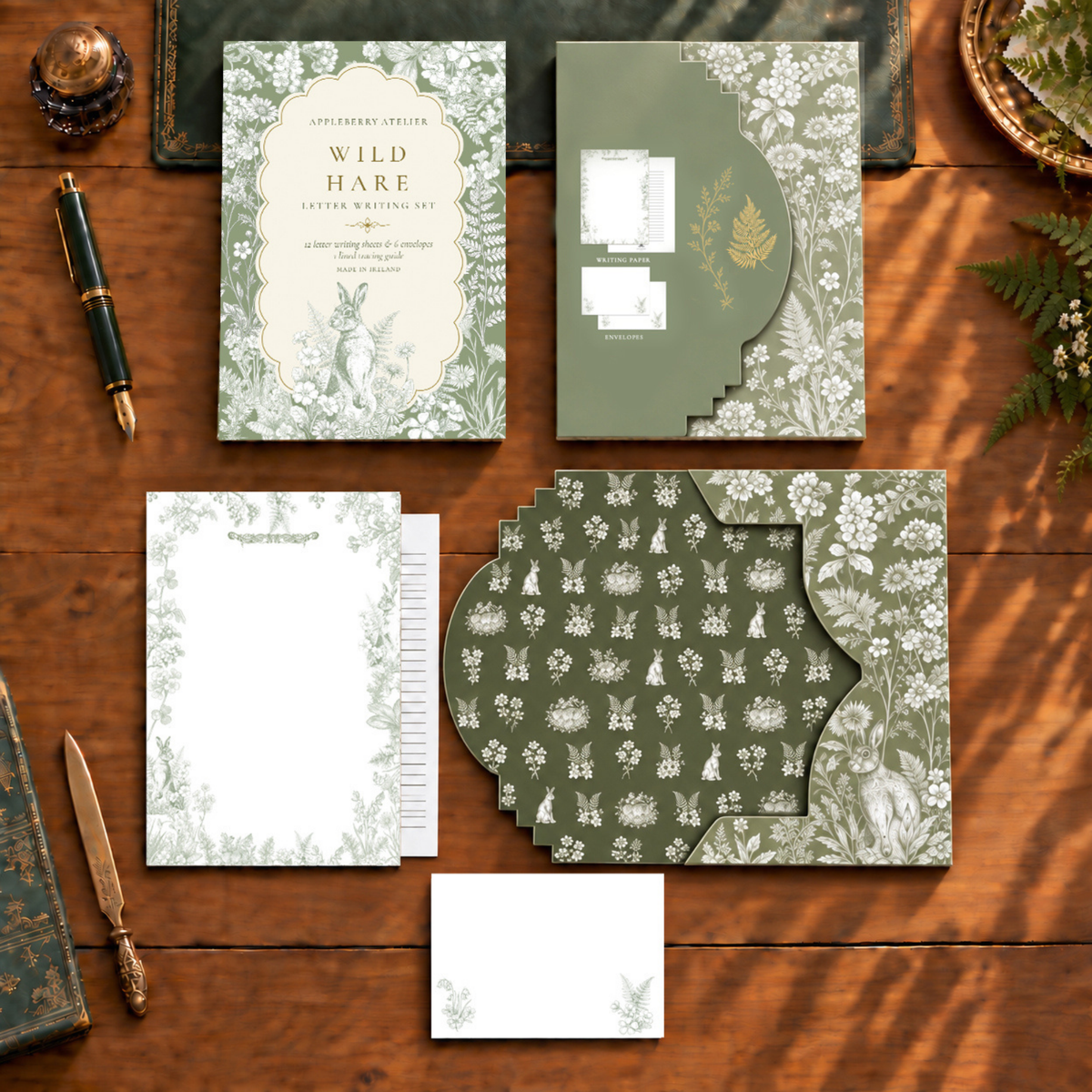 A coordinated stationery set arranged on a wooden tabletop, including a storage envelope with a rabbit illustration and the words 'Wild Hare Letter Envelope,' matching writing paper and envelope. The design uses muted sage green, cream, and white floral/rabbit motifs, with a few gold-toned decorative accents.