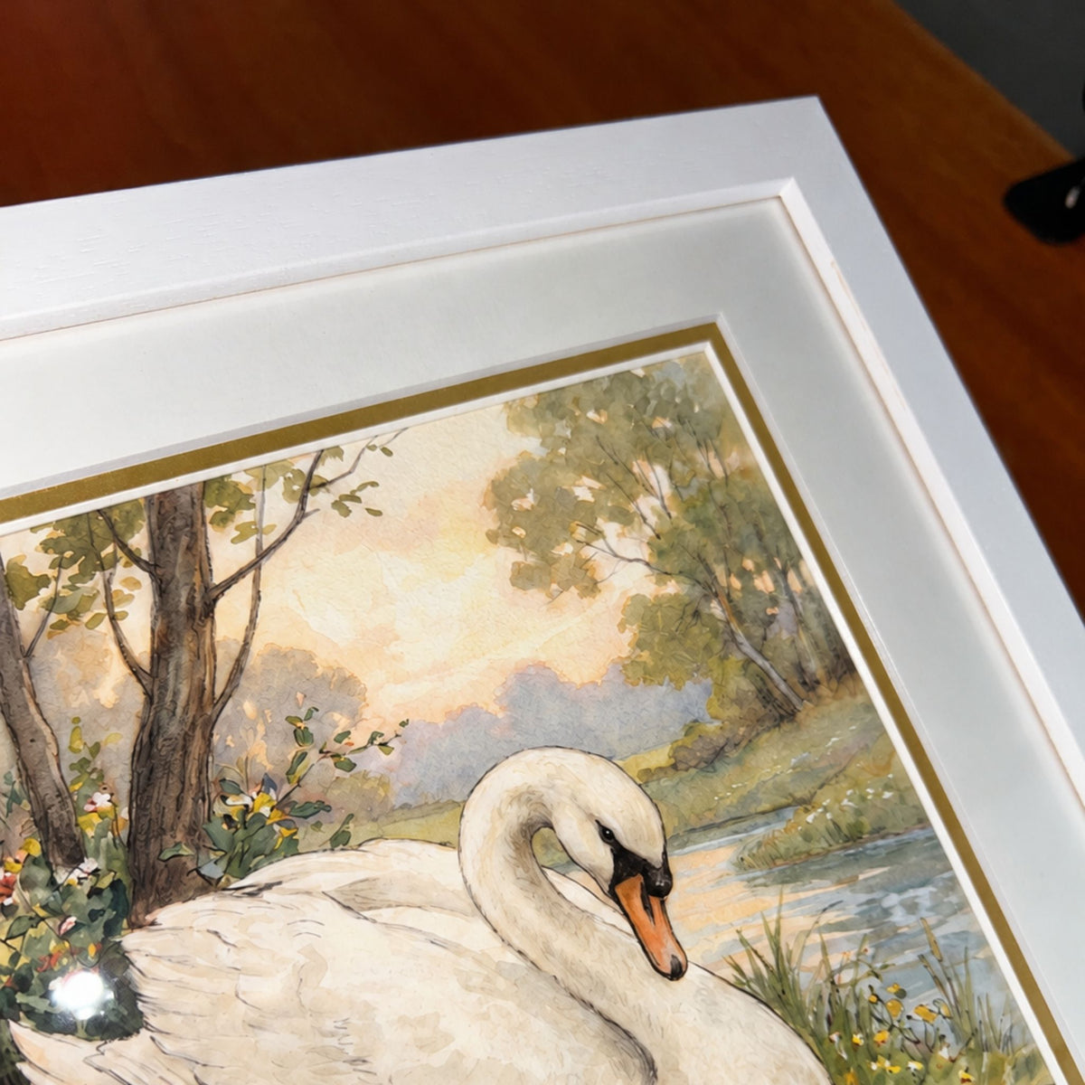 A framed art print featuring a white swan in a scenic landscape with trees, water, and soft pastel sky, shown at an angle on a wooden surface.