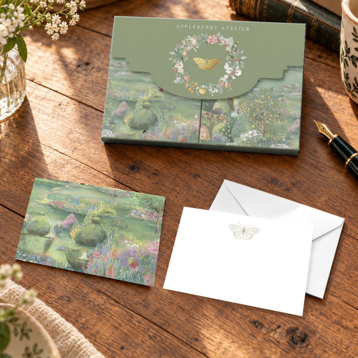 A styled flat-lay of a stationery set on a wooden table, featuring a green illustrated envelope box with a butterfly and floral wreath design, a matching illustrated notelet, and white envelopes.
