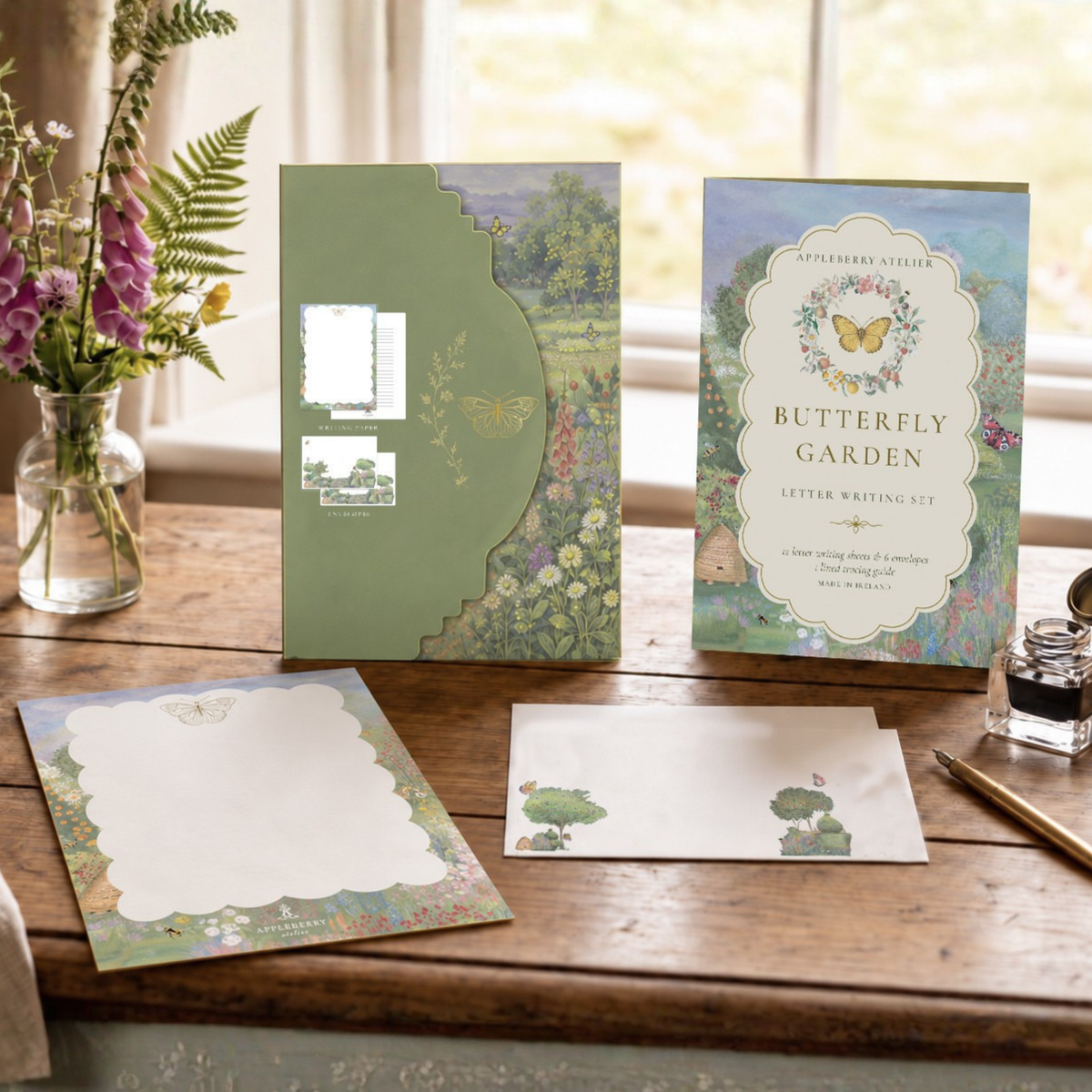 A butterfly garden letter writing set displayed on a wooden table by a window, including a green storage folder, a floral card, a letter, an envelope, and a writing kit box.
