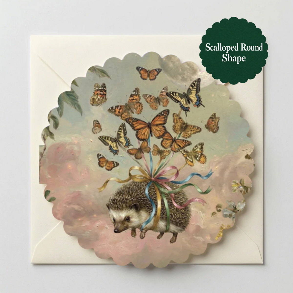 A scalloped round greeting card featuring an illustration of a hedgehog tied with a rainbow ribbon, surrounded by butterflies in soft pink and blue tones, shown on a white envelope against a light gray background. A dark green badge in the top right reads 'Scalloped Round Shape'.