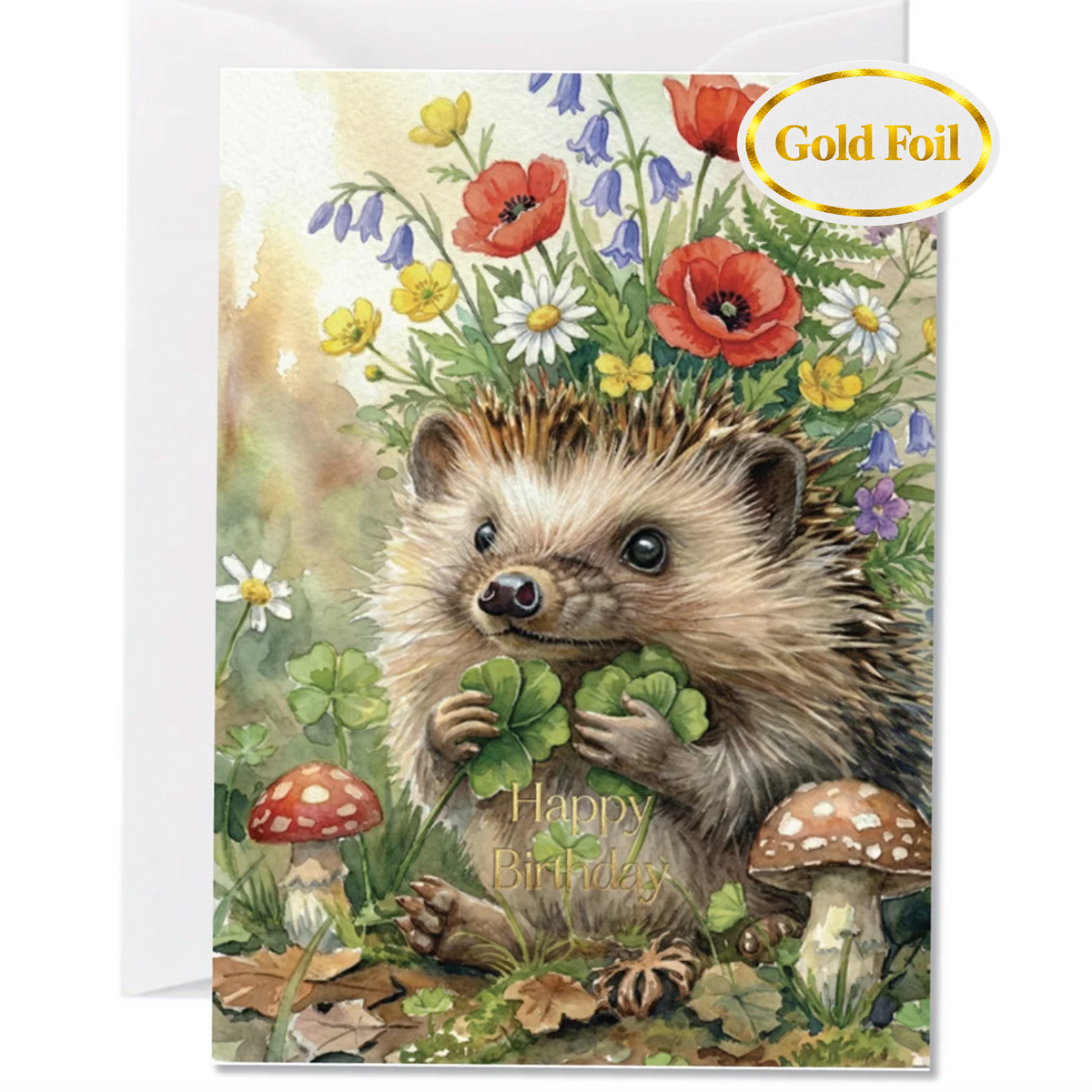 A square greeting card featuring a painted hedgehog holding clovers in a flower-filled meadow, with the words 'Happy Birthday' on the card. A gold foil stamp is visible in the top right corner, and a white envelope is partially visible behind the card.