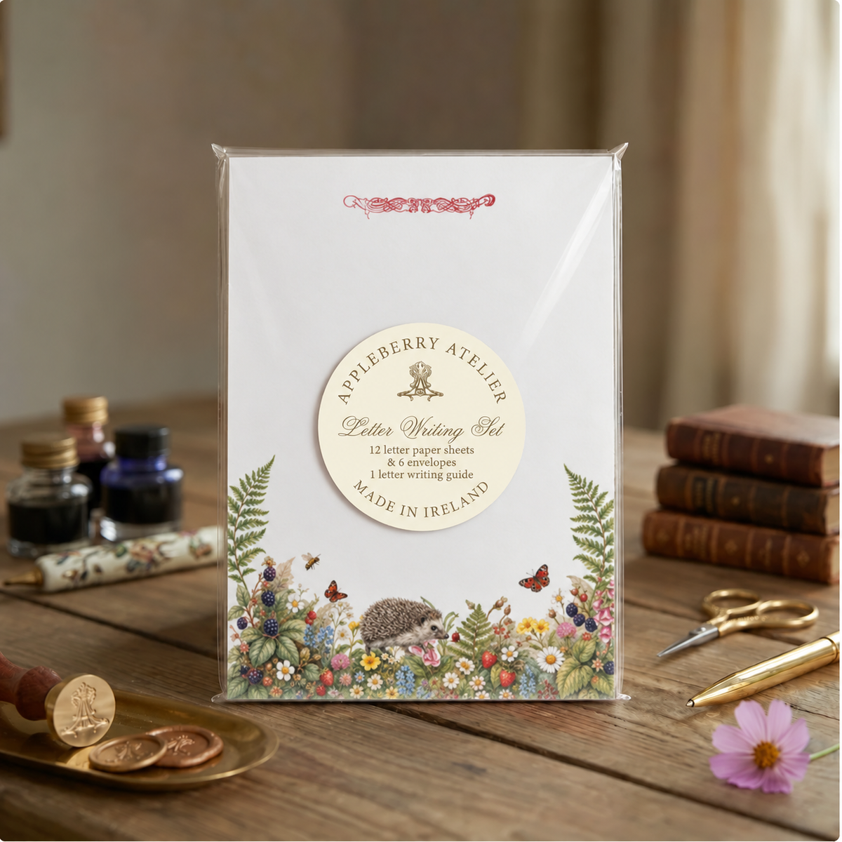 A letter-writing set stands on a wooden table in a styled indoor scene. The package is clear plastic-wrapped and shows a white front with a round cream label reading 'Appleberry Atelier Letter Writing Set' and floral/animal illustrations along the bottom.