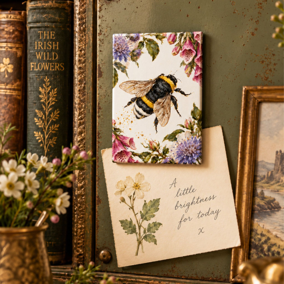A small rectangular floral Magnet featuring a bumblebee and purple flowers, styled on a vintage green surface with an old book, framed picture, and a card reading 'A little brightness for today'.