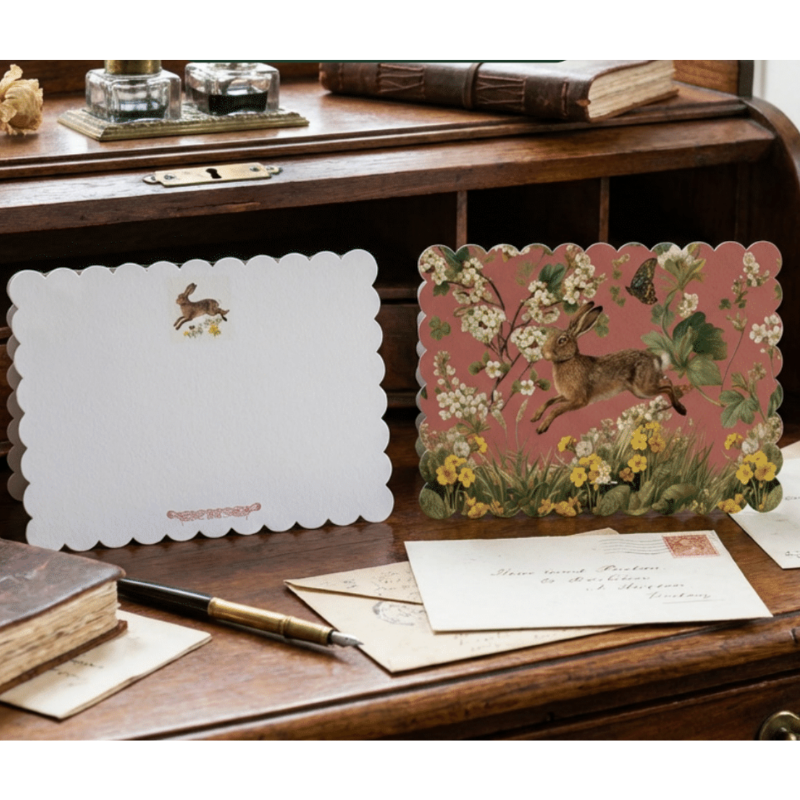 A styled flat-lay of vintage stationery on a wooden desk, featuring a white scalloped note card with a small deer illustration and a pink floral greeting card with a rabbit and flowers.