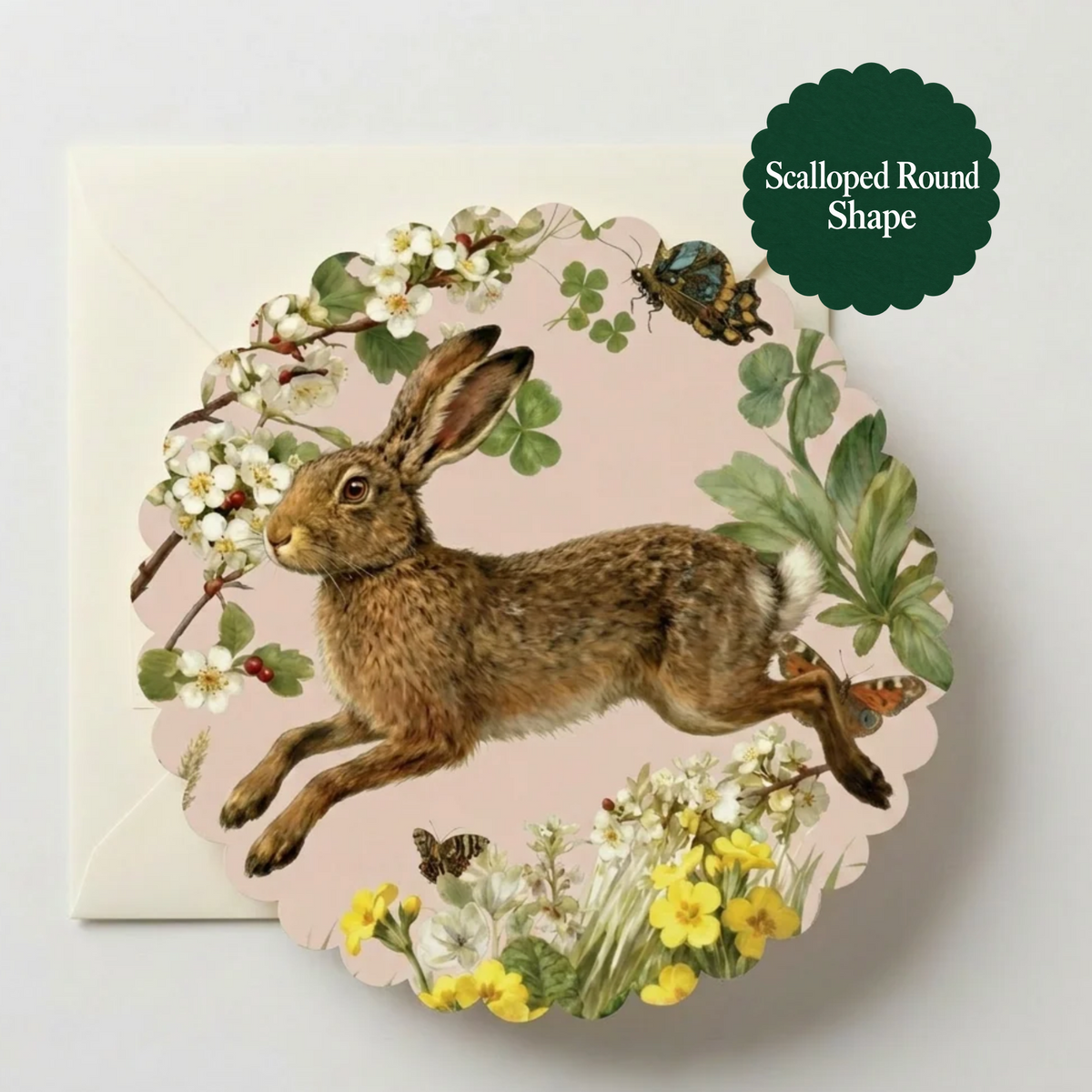 A scalloped round greeting card featuring an illustration of a brown hare among white blossoms, green leaves, yellow flowers, and butterflies, shown with a white envelope. A dark green badge in the top right reads 'Scalloped Round Shape'.