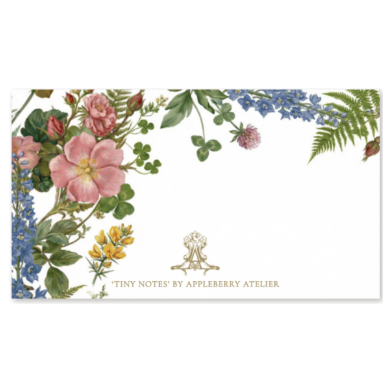 A rectangular white stationery card or note pad with a botanical border of pink flowers, blue blossoms, green leaves, and ferns along the top and left edges. Gold text at the bottom reads “TINY NOTES’ BY APPLEBERRY ATELIER,” with a small gold crest above it.